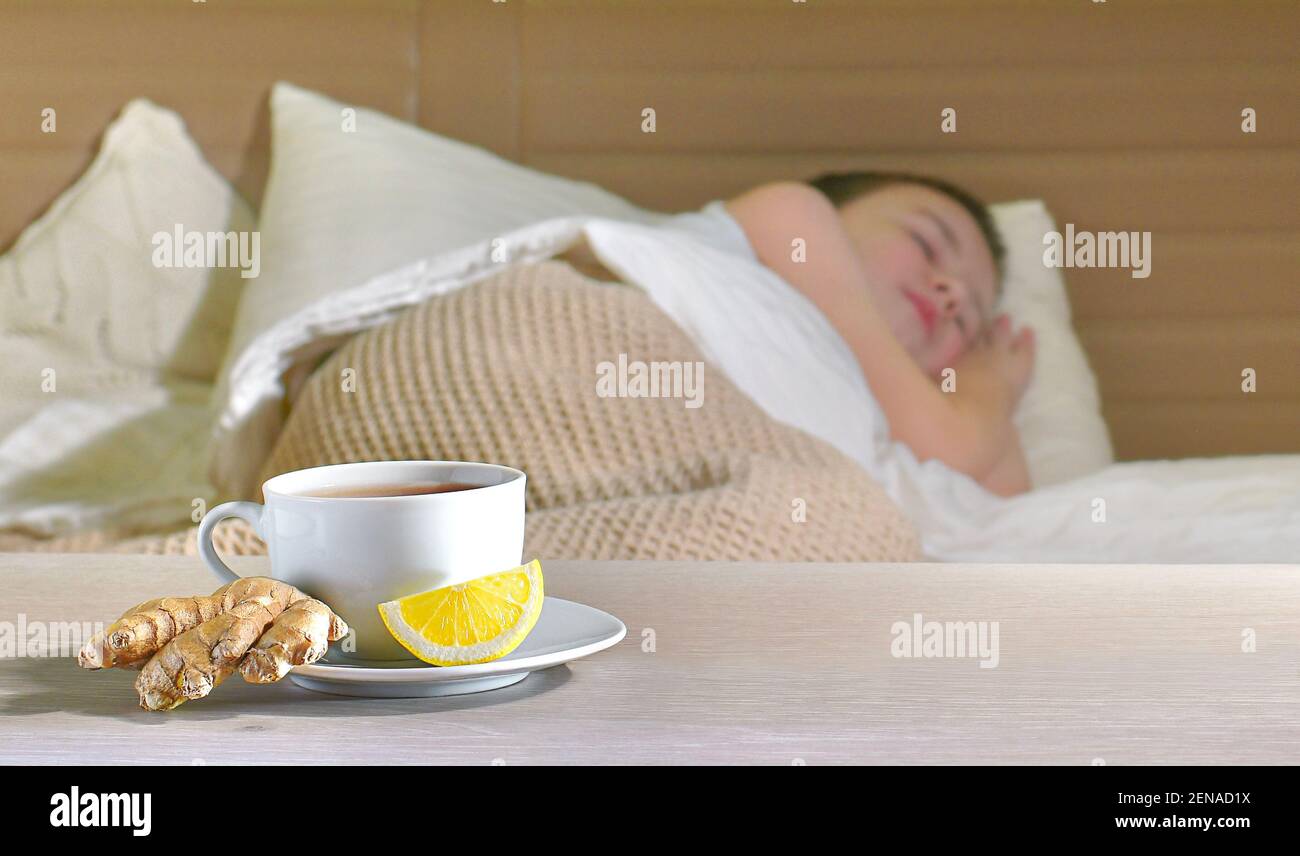 Una tazza di tè su un piatto con radice di zenzero e una fetta di limone su sfondo sfocato. Dormire bambino malato in letto. Foto Stock