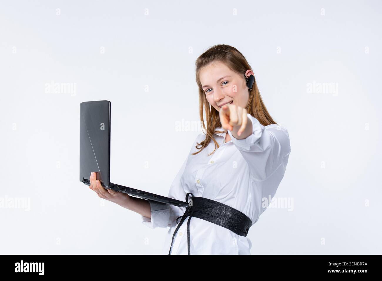 Attraente giovane donna caucasica si erge su uno sfondo grigio isolato tiene un laptop guarda direttamente con sorriso amichevole e punti mano alla fotocamera Foto Stock