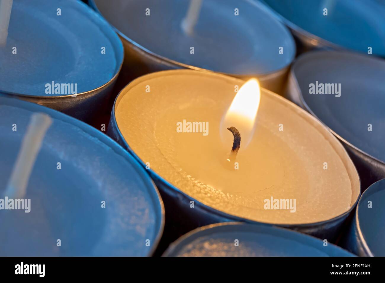 Primo piano delle luci del tè con una accesa. Una sola fiamma arancione calda tra le candele in ombra blu. Natale, condoglianze o funerali. Foto Stock