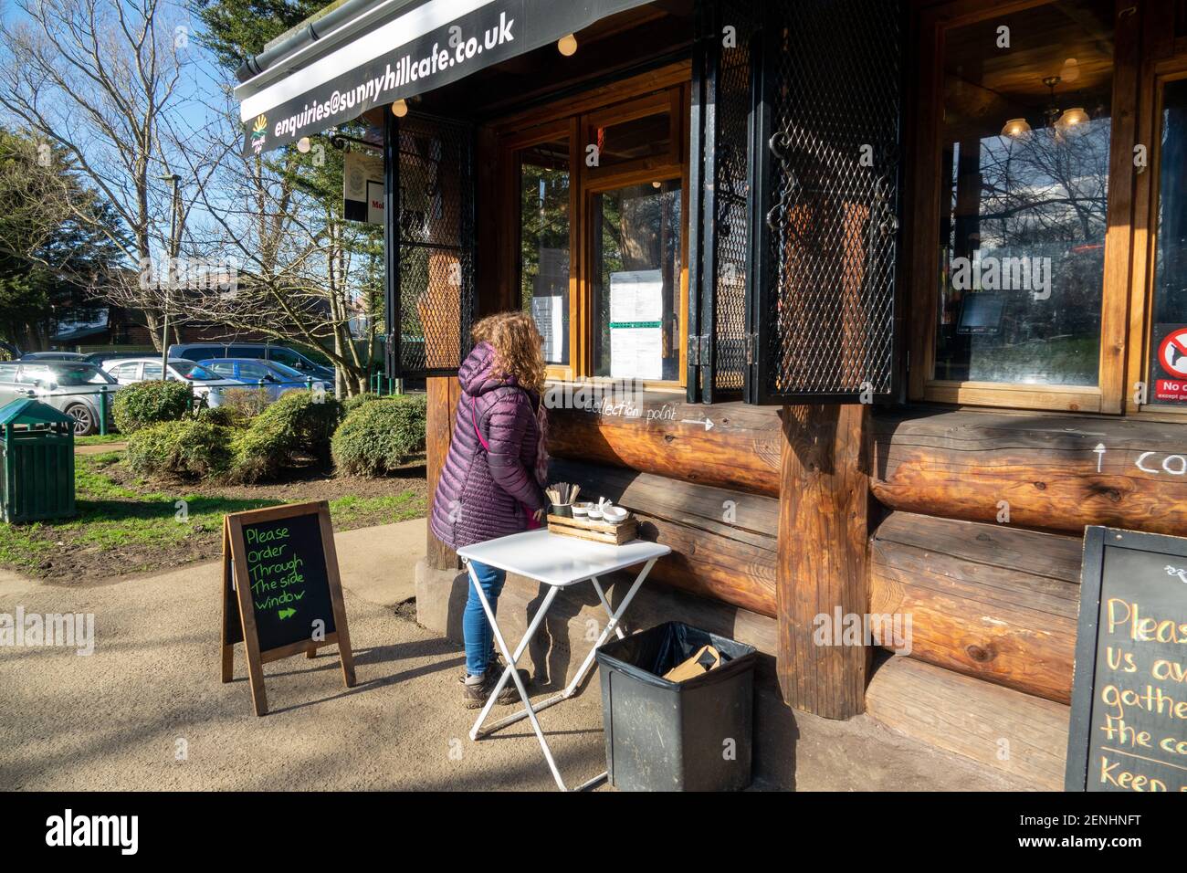 Donna che ordina un drink e un cibo in un locale all'aperto. Code socialmente distanziate di persone che ordinano bevande al Sunny Hill Cafe, una caffetteria a Sunny Hill Park, Hendon. Inizio di un weekend soleggiato con cielo blu.. Regno Unito Meteo. Londra, Regno Unito. 26 febbraio 2021. Foto Stock