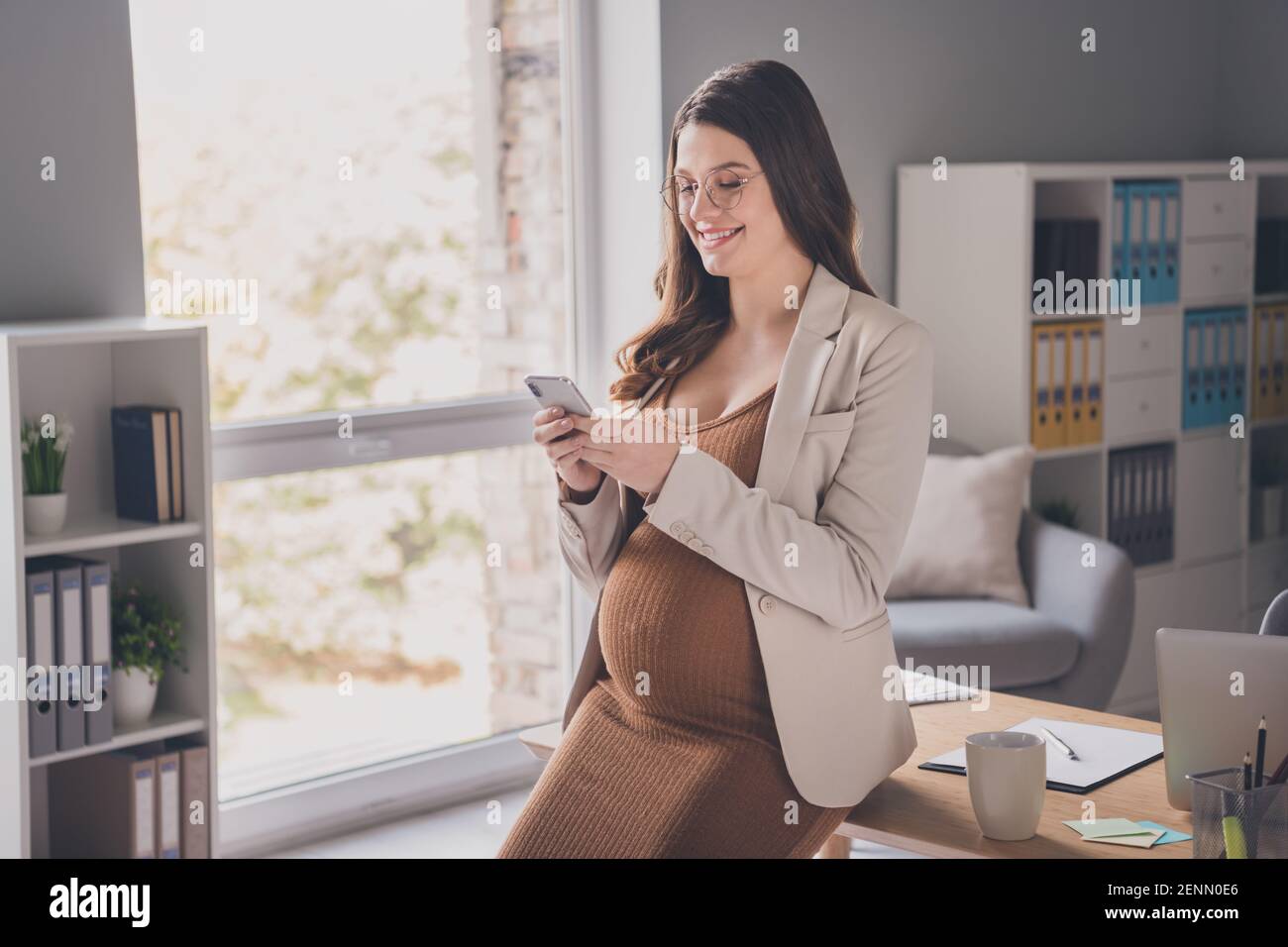 Ritratto fotografico di una donna incinta che tiene il telefono in due mani navigazione seduta sulla scrivania in un ufficio moderno Foto Stock