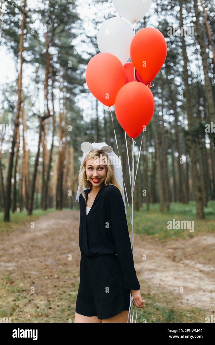 Felice ragazza in velo sposa e balloni bianchi e rossi in piedi nel parco e sorridente. Ragazza sulla festa di gallina Foto Stock