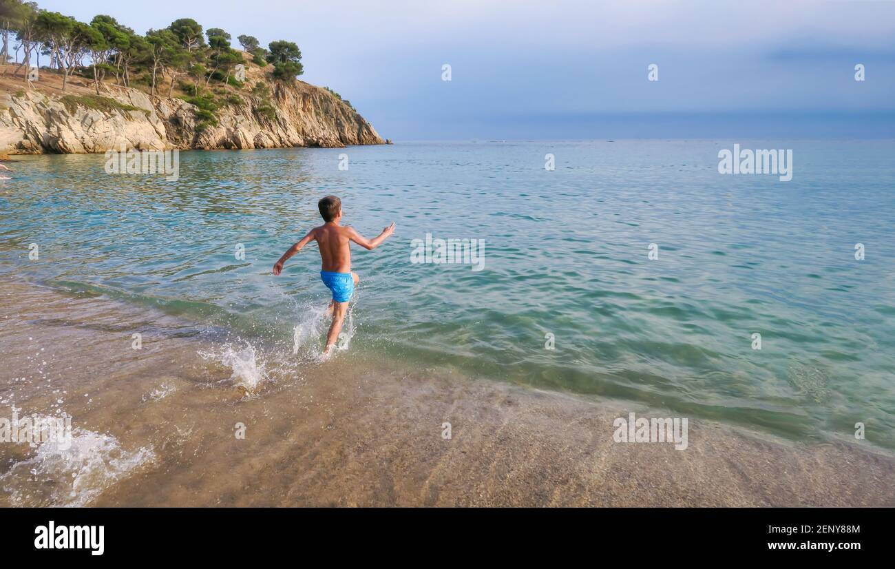 il bambino corre lungo la riva verso il mare spruzzi d'acqua per fare un tuffo in una spiaggia di cristallo chiaro acque turchesi con una scogliera sullo sfondo o Foto Stock