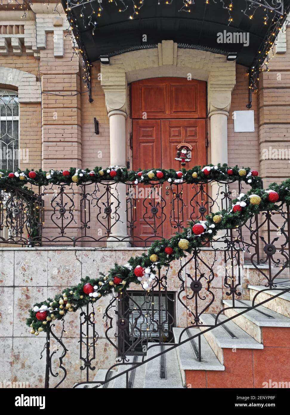 Idea decorazione portico di Natale. Ingresso casa con porta rossa decorata  per le vacanze. Corona rossa e verde di rami di abete e luci sulla  ringhiera. Vigilia di Natale a casa Foto