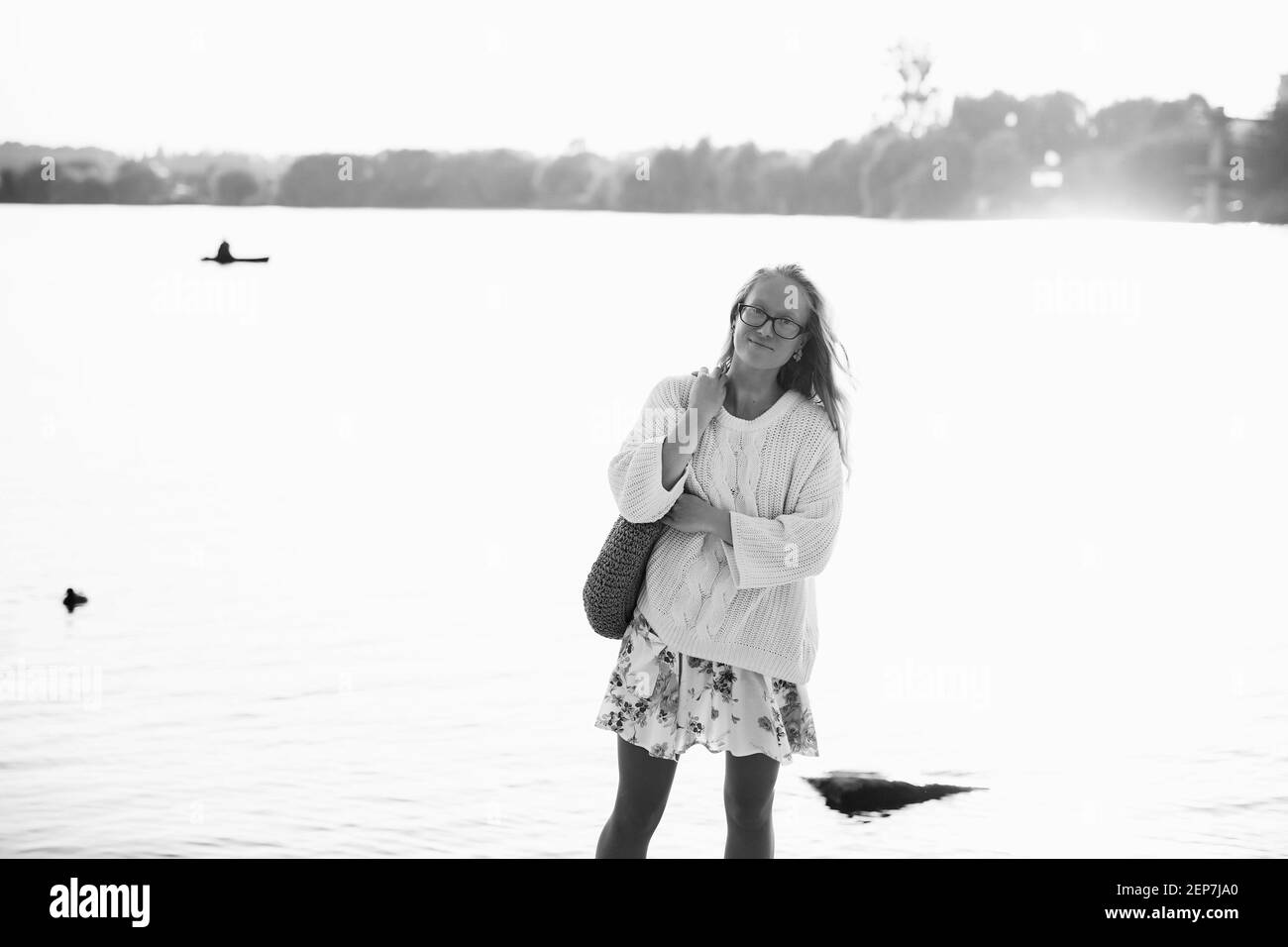 Giovane bella bionda, bella donna, ragazza che cammina sulla spiaggia in un vestito e maglione con fiori in una giornata di sole vicino al lago, fiume Foto Stock