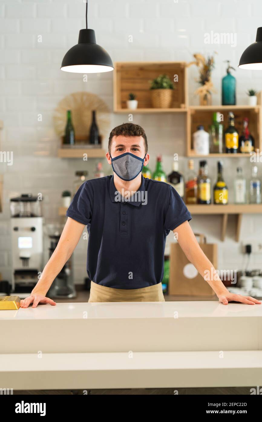 ritratto di un cameriere professionista con maschera in posa dietro il bar di un ristorante durante la pandemia di coronavirus Foto Stock
