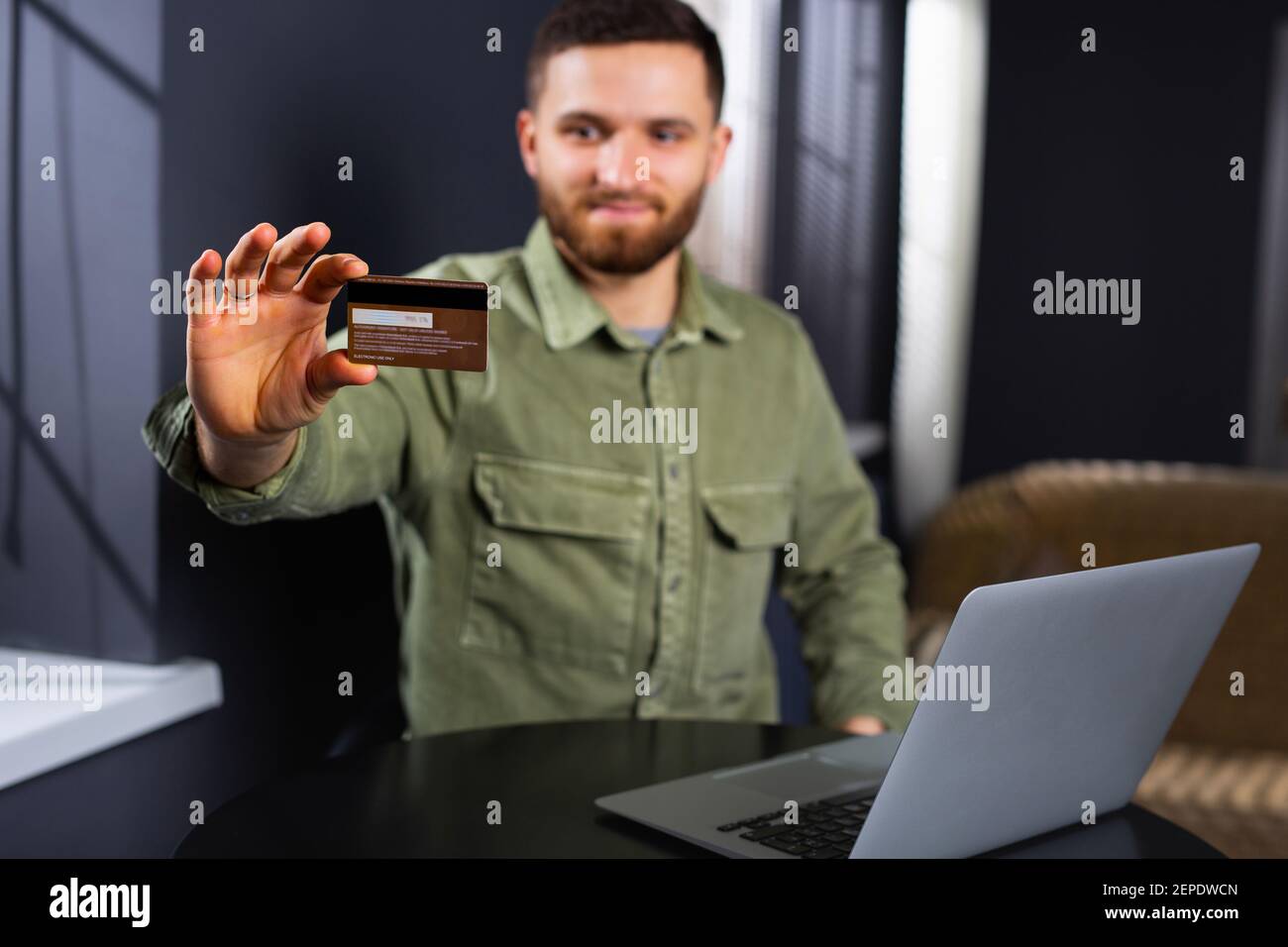 Una foto sfocata di un uomo seduto davanti a un computer portatile e che tiene fuori una carta di plastica della banca alla fotocamera. Prestiti e acquisti online. Foto Stock