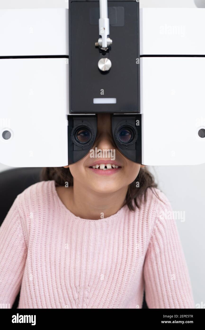 Faccia della ragazza carina felice che siede davanti oftalmologico attrezzatura mentre la sua vista ha controllato nelle cliniche moderne Foto Stock