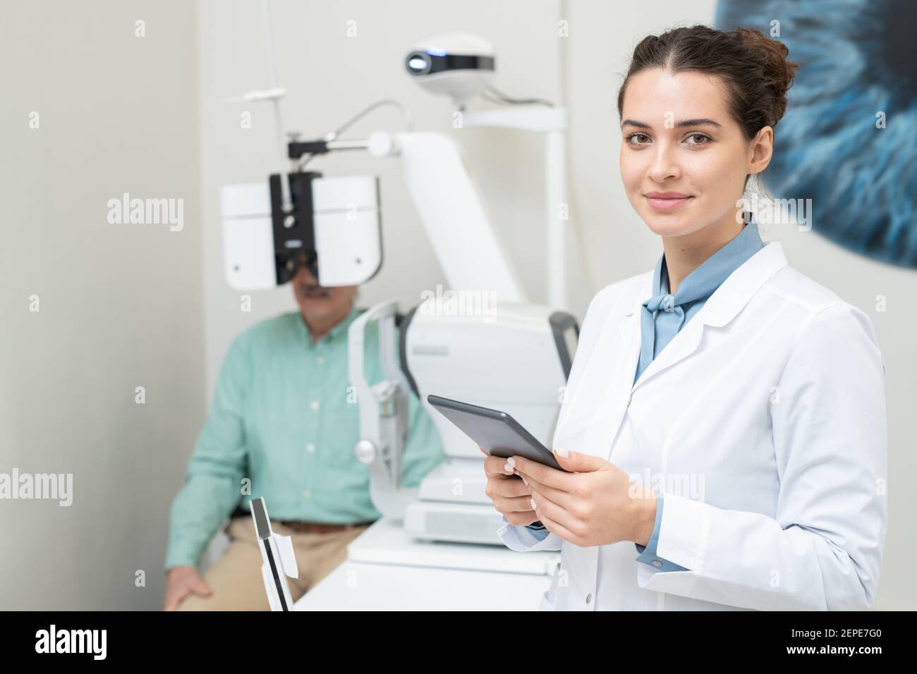 Felice giovane oftalmologo femminile in whitecoat che ti guarda mentre uso della compressa sullo sfondo del paziente con la sua vista controllata su Foto Stock