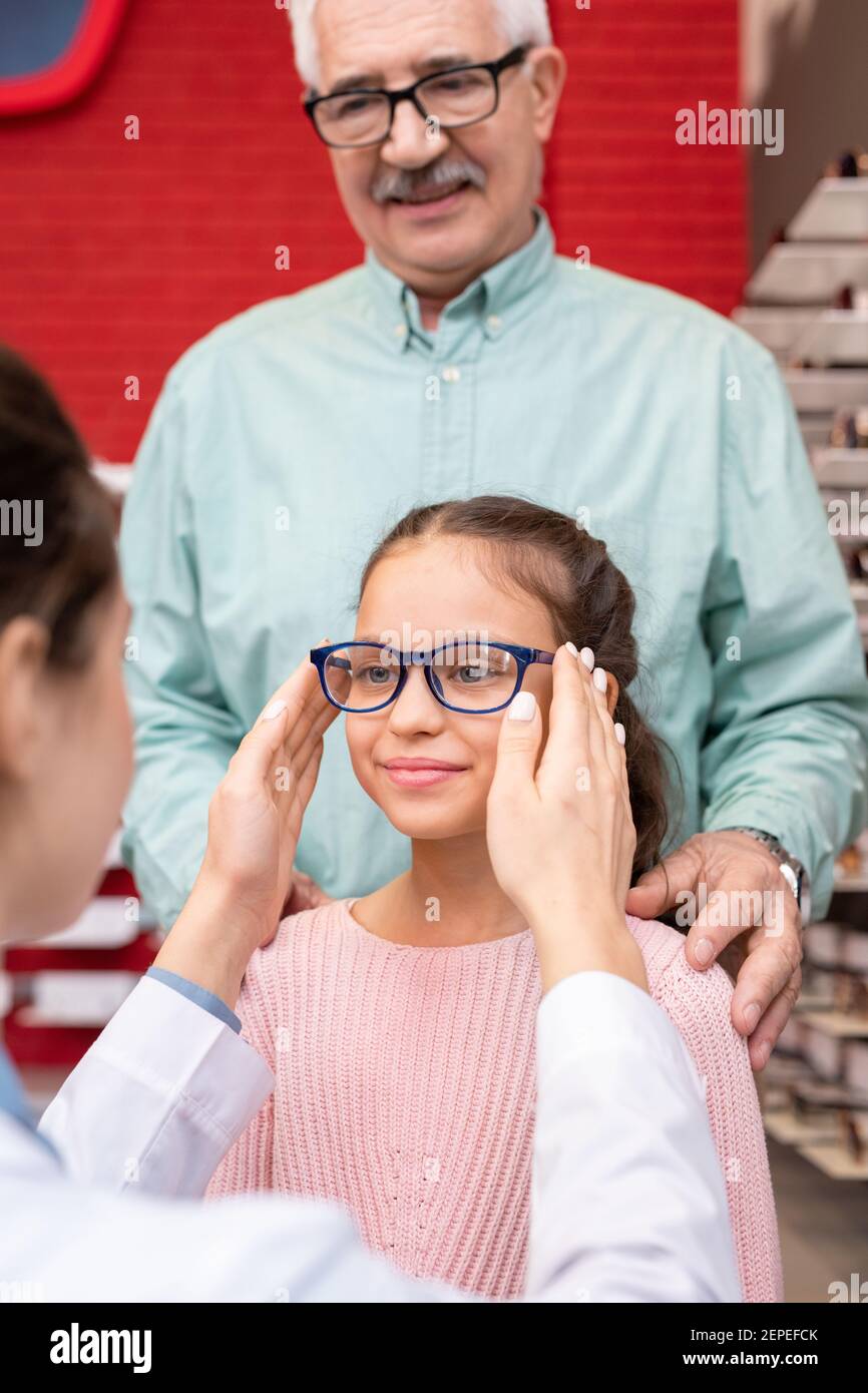 Giovane ottico femminile che mette un paio di nuovi occhiali sul viso di carino piccola ragazza che visita ufficio oftalmologico o cliniche con suo nonno Foto Stock