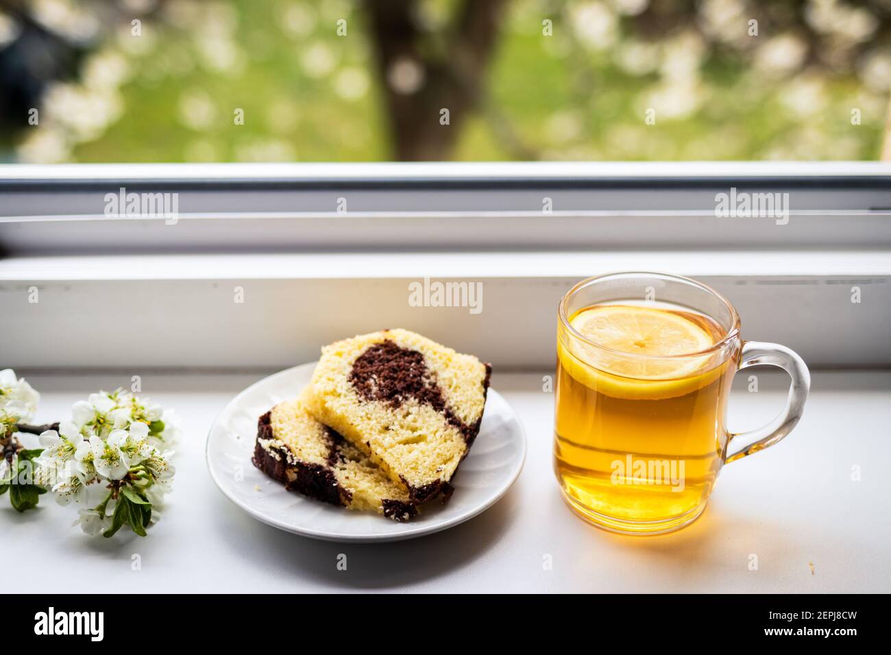 Tazza da tè con limone, fetta di torta e fiori su parapetto. Vista dalla finestra all'albero in fiore. Godimento della prima colazione alla mattina di primavera Foto Stock