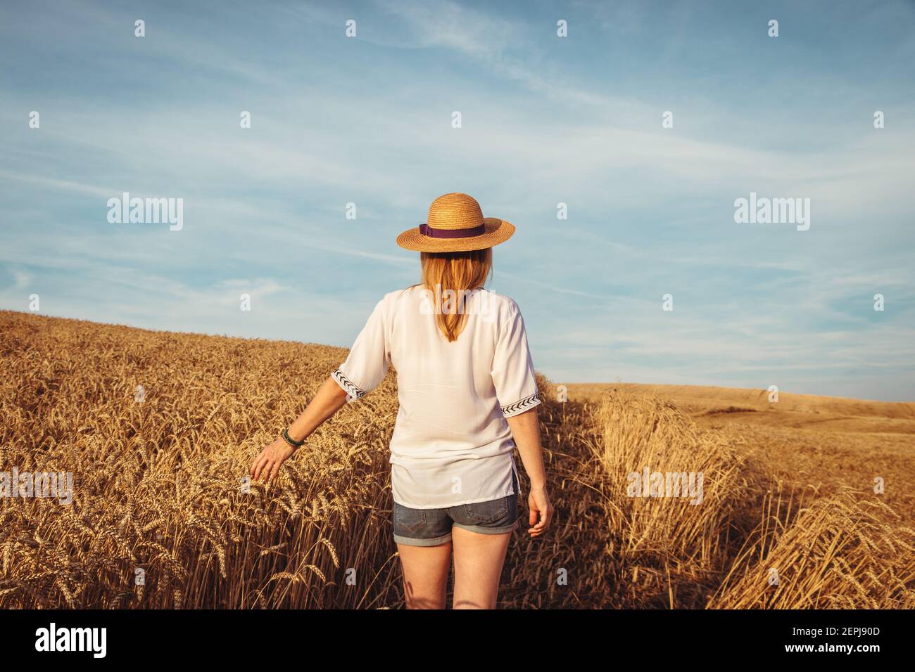 Donna contadina in piedi sul campo. Godimento di estate in campagna. Capelli biondi donna caucasica che indossa cappello di paglia, camicia bianca e pantaloncini. Foto Stock