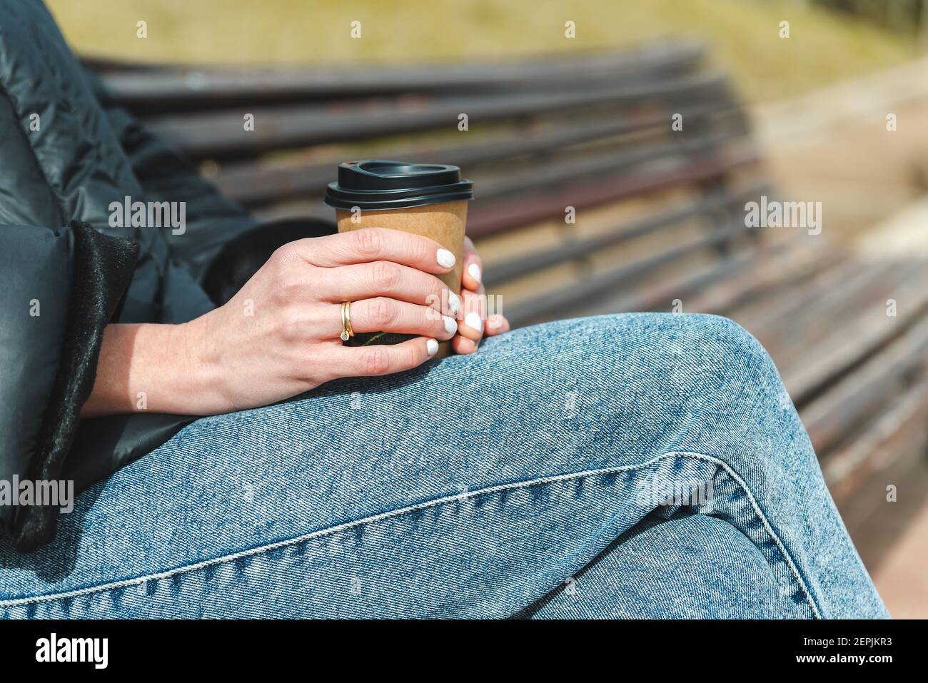 primo piano prendere le mani di una donna bianca che tiene una tazza di carta usa e getta di caffè. Indossa jeans blu, cappotto nero e un anello sulle dita. È seduta su una panchina di legno in un parco verde urbano Foto Stock