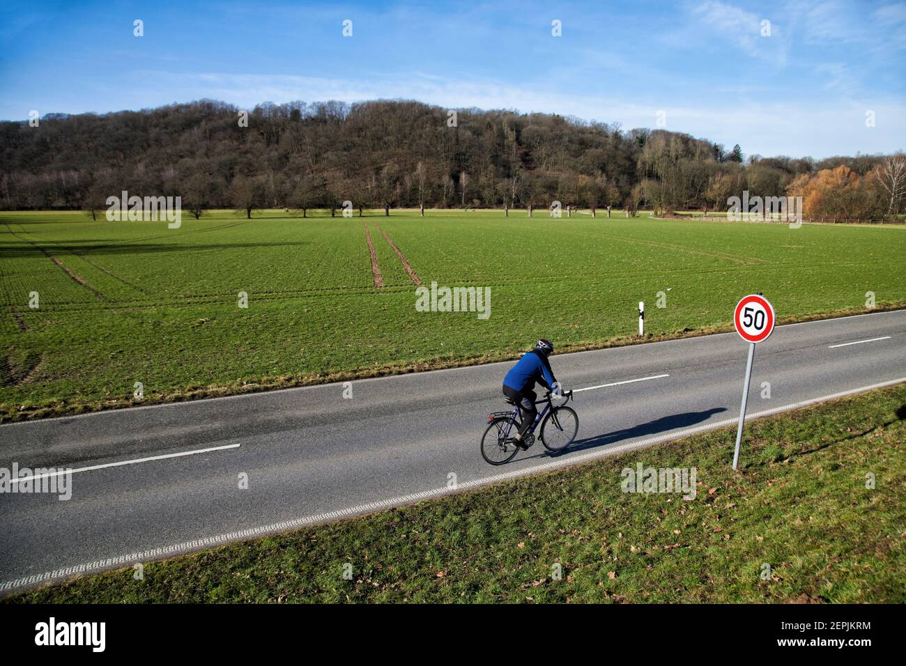 Fahrradfahrer auf einer zweispurigen Landstrasse vor einem Feld. Im Hintergrund befindet sich ein bewaldeter Huegel. Es ist Inverno. Zu sehen ist ein V Foto Stock