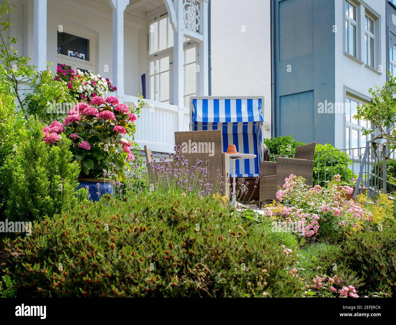Tipico cortile di fronte ad una casa di legno bianco con una sedia da spiaggia di vimini blu. Il giardino è molto verde e colorato con belle piante verdi. Foto Stock