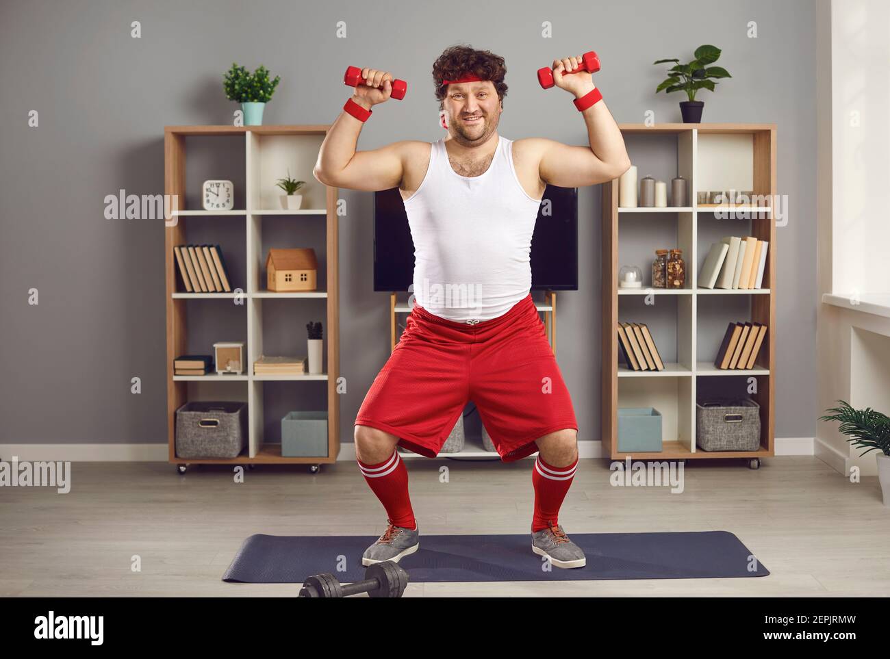 Divertente uomo chubby che si esercita con manubri in piedi su tappetino fitness in soggiorno Foto Stock