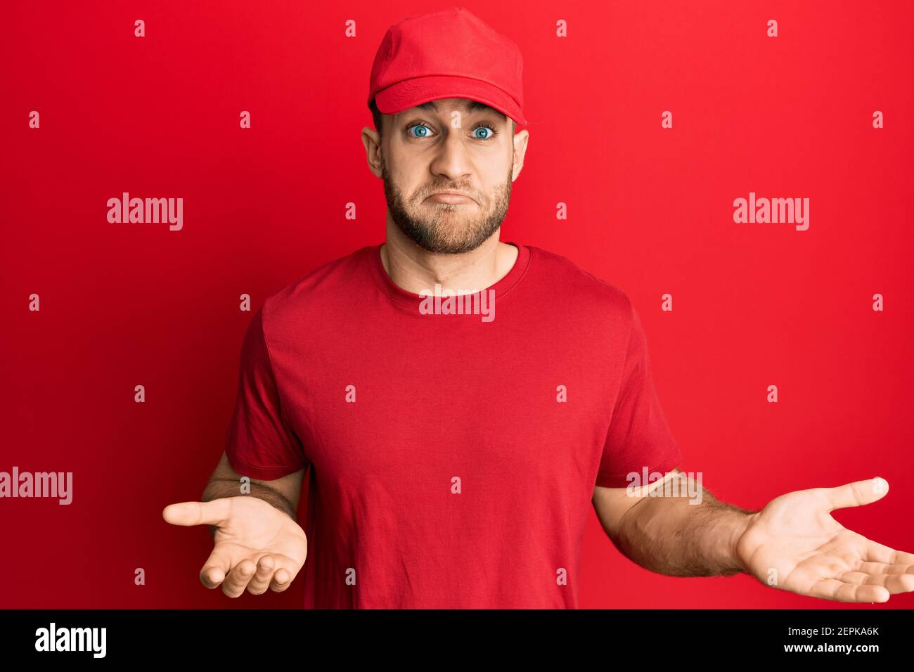 Giovane uomo caucasico che indossa uniforme di consegna e cappello indecore e confuso espressione con braccia e mani sollevate. Concetto di dubbio. Foto Stock