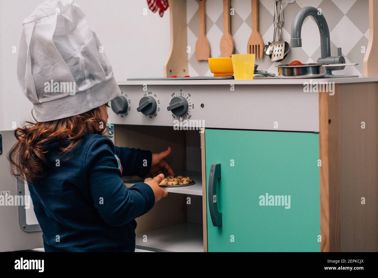 Bambina caucasica, indossando un cappello da chef, giocando ad essere una cuoca nella sua piccola cucina giocattolo. Foto Stock