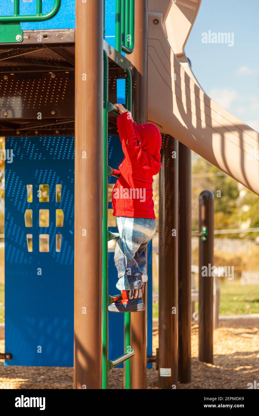 Un bambino bianco sta giocando in un parco giochi da solo. Sta tenendo le barre di una scala metallica e sta cercando di tirarsi in su mentre impara a salire. Su Foto Stock