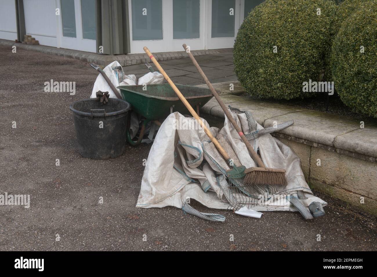 Old Rusty Wheelbarrow con un pennello e rastrello su un sentiero in un giardino nel Devon rurale, Inghilterra, Regno Unito Foto Stock