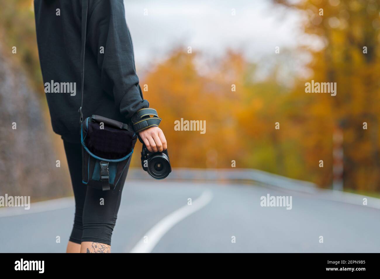 Vista posteriore fotografo anonimo in abbigliamento casual in piedi con professionista fotocamera su strada rurale vuota che attraversa la foresta gialla in autunno da Foto Stock
