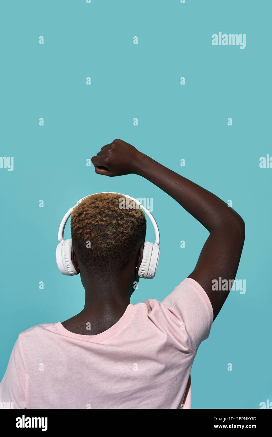 Vista posteriore della donna afro-americana senza volto con braccio rialzato e ha chiuso l'ascolto della musica con le cuffie mentre si è in piedi su sfondo blu Foto Stock