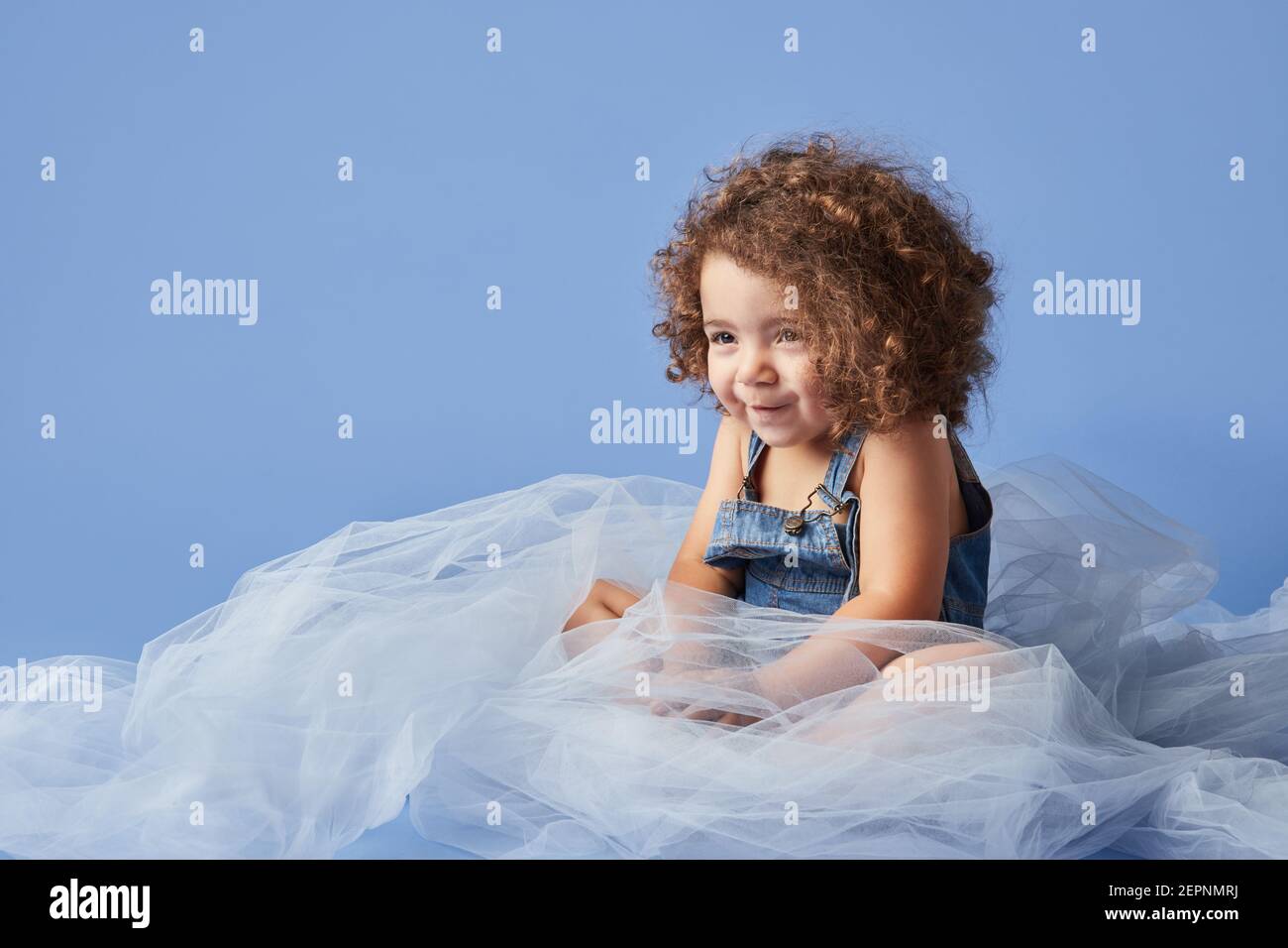 Adorabile ragazza dai capelli ricci sorridente carino seduta su tessuto sottile su sfondo blu e guardando via Foto Stock