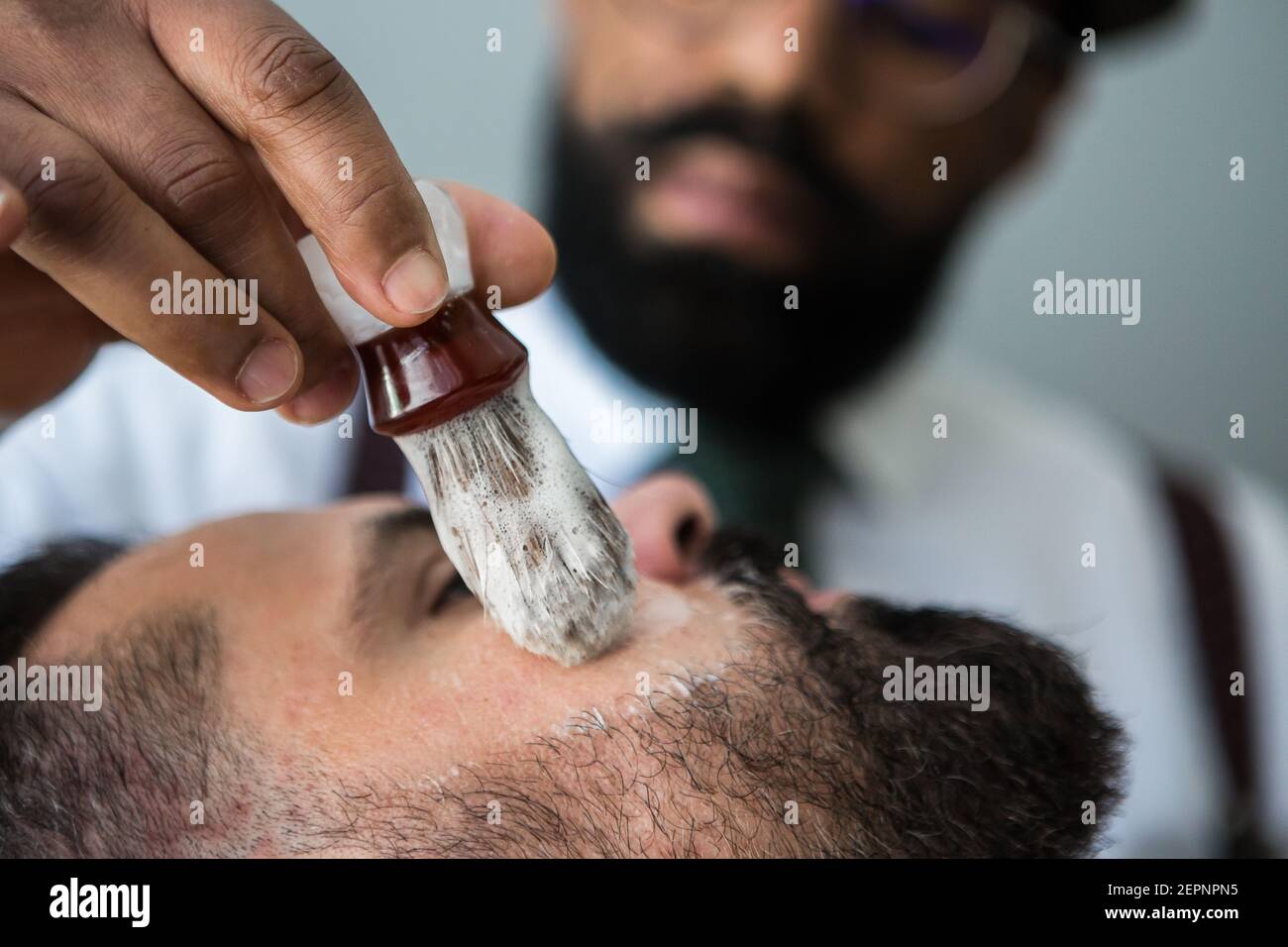 Primo piano di crop maschile etnico beauty master applicando una rasatura liscia sapone sulla faccia di uomo bearded usando la spazzola in acconciatura salone Foto Stock