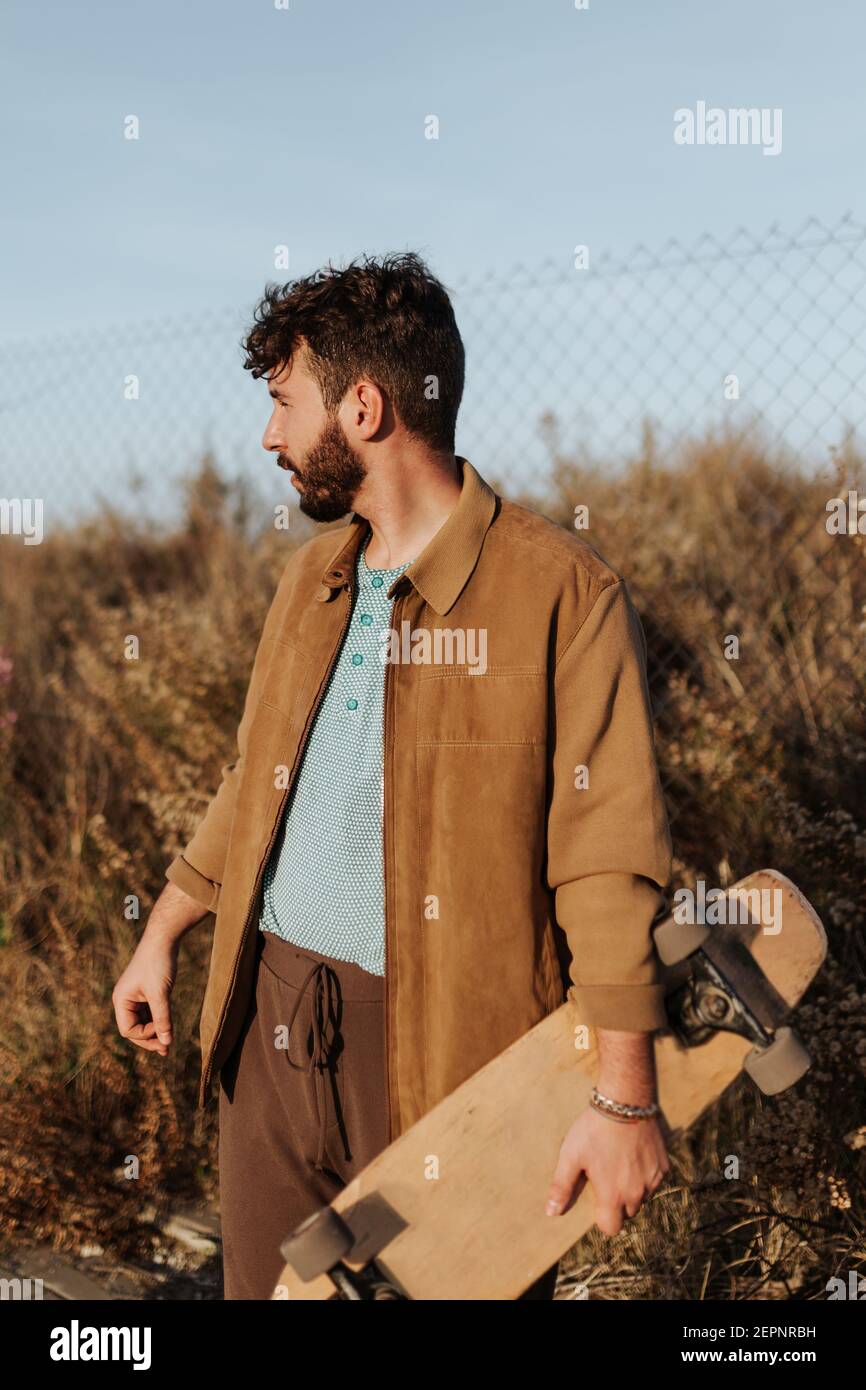 Giovane skater maschile bearded in abbigliamento casual in piedi vicino all'erba e recinzione con skateboard che guarda lontano Foto Stock