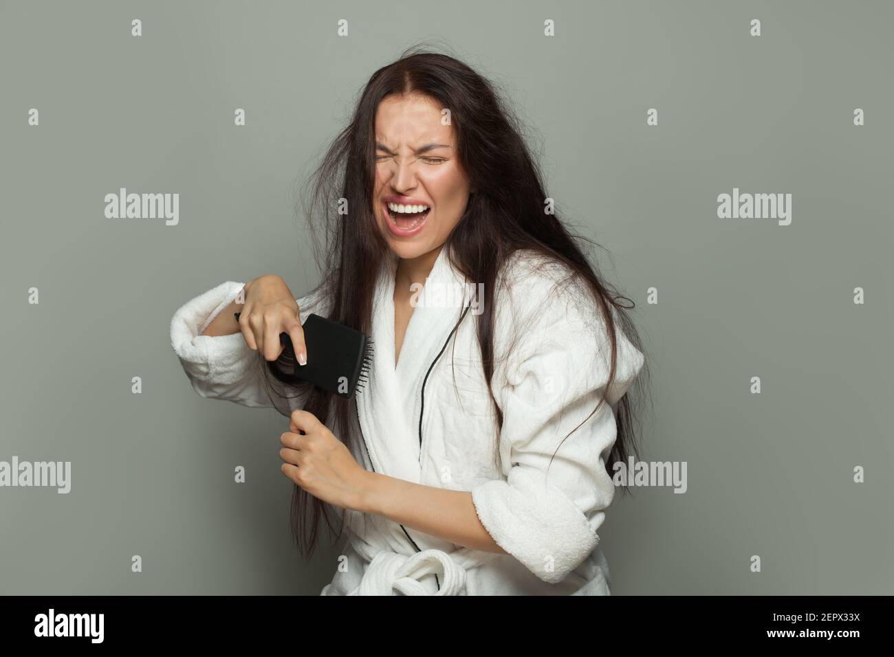 Donna arrabbiata che combatte i suoi capelli aggrovigliati su sfondo bianco. Cura dei capelli e concetto di problema dei capelli Foto Stock