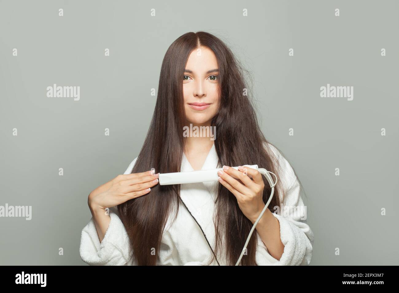 Perfetta donna brunetta raddrizza i capelli con la piastra su sfondo bianco. Concetto di cura dei capelli Foto Stock