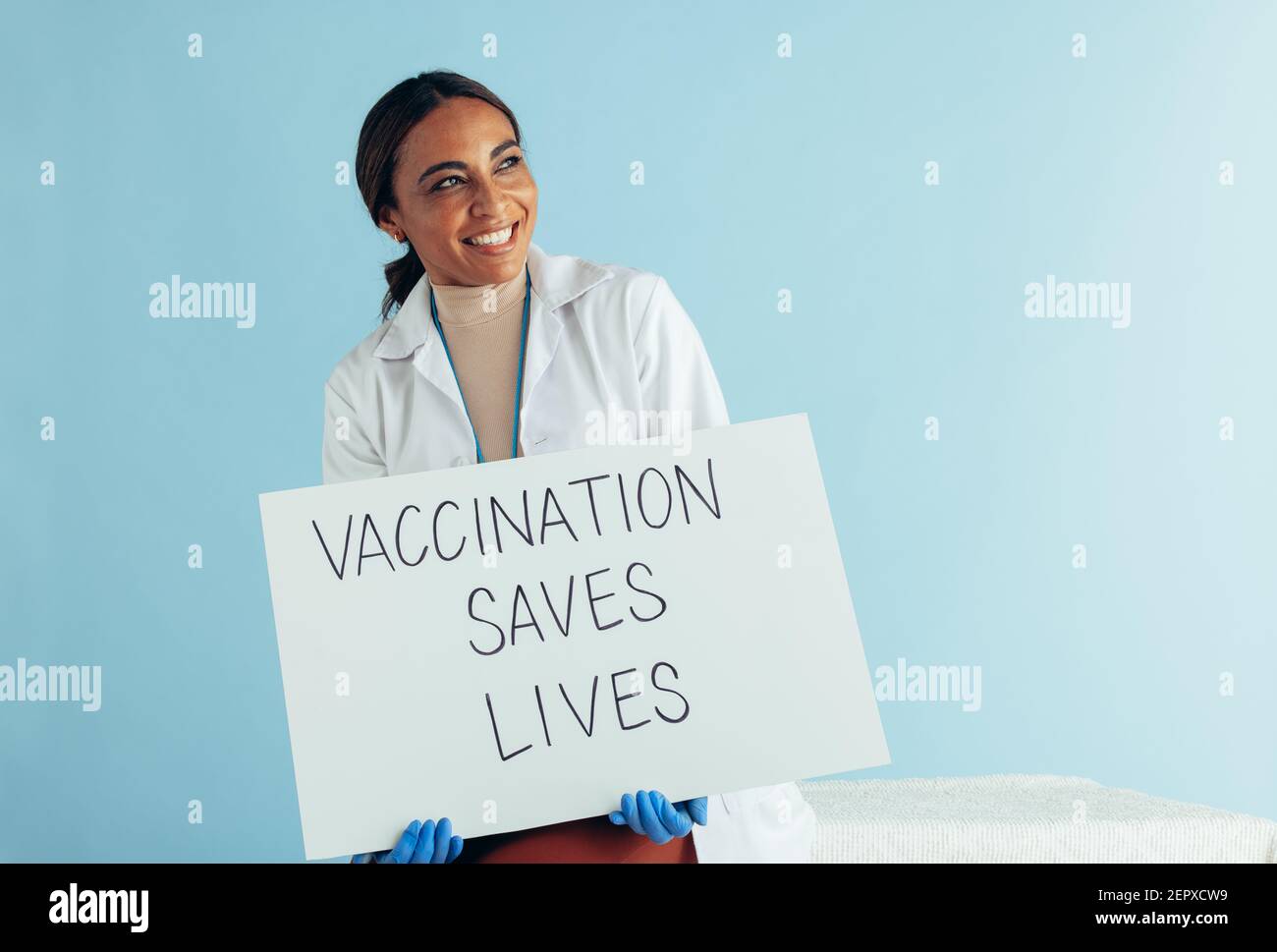 Femmina medico che tiene bandiera di 'la vaccinazione salva la vita' e guardando via sorridente. Medico con un cartello su sfondo blu. Foto Stock