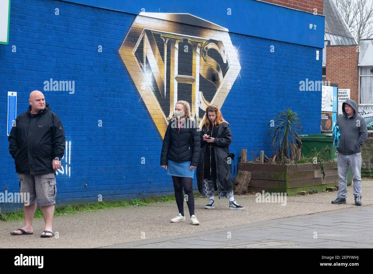 Londra, UK, 28 febbraio 2021: Un murale a Tulse Hill raffigura il logo NHS nello stile di un badge supereroe. Passers-by prendere l'esercizio o la coda per il negozio Co-op dietro l'angolo, alcuni indossando maschere viso e tutti socialmente distanziando. Anna Watson/Alamy Live News Foto Stock