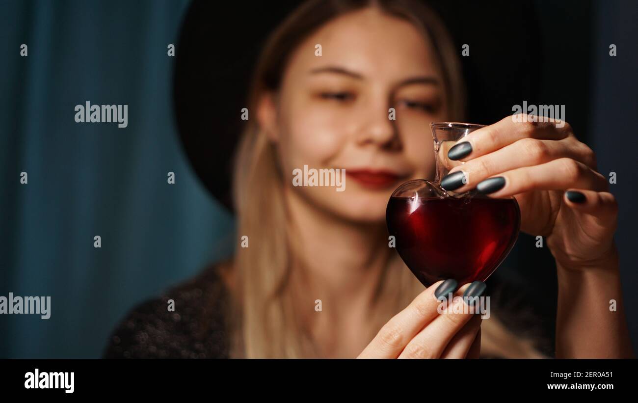 Immagine tagliata di donna che tiene il vaso di vetro a forma di cuore di pozione d'amore. Bella giovane donna in un cappello di strega. Mettere a fuoco su Potion o Poison Vial Foto Stock