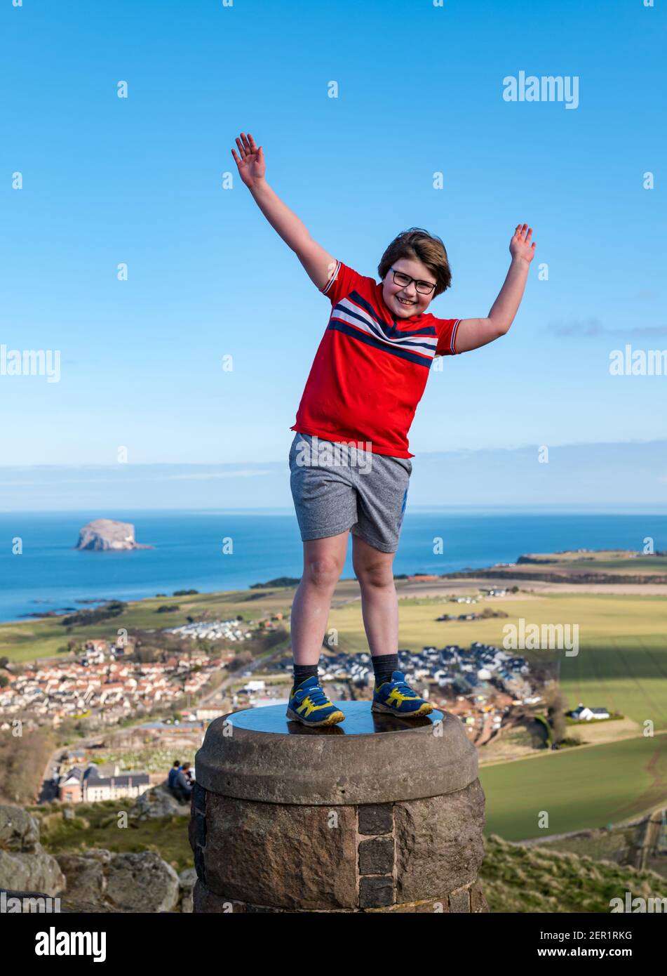 North Berwick, East Lothian, Scozia, Regno Unito, 28 febbraio 2021. Arrampicata Berwick Legge per la sindrome di Rett: Sulla malattia rara giorno di 7 anni Nina McKinney finisce il suo obiettivo di salire la collina di 613ft ogni giorno questo mese per raccogliere soldi per Reverse Rett, una carità che sostiene la ricerca nel disordine di cui la sorella soffre. Nella foto: Nina celebra la fine della sua campagna di raccolta fondi in cima alla collina dopo aver sfidato tutte le condizioni meteorologiche durante il mese in piedi su un indicatore di punto di vista con Bass Rock nel Firth of Forth sullo sfondo Foto Stock