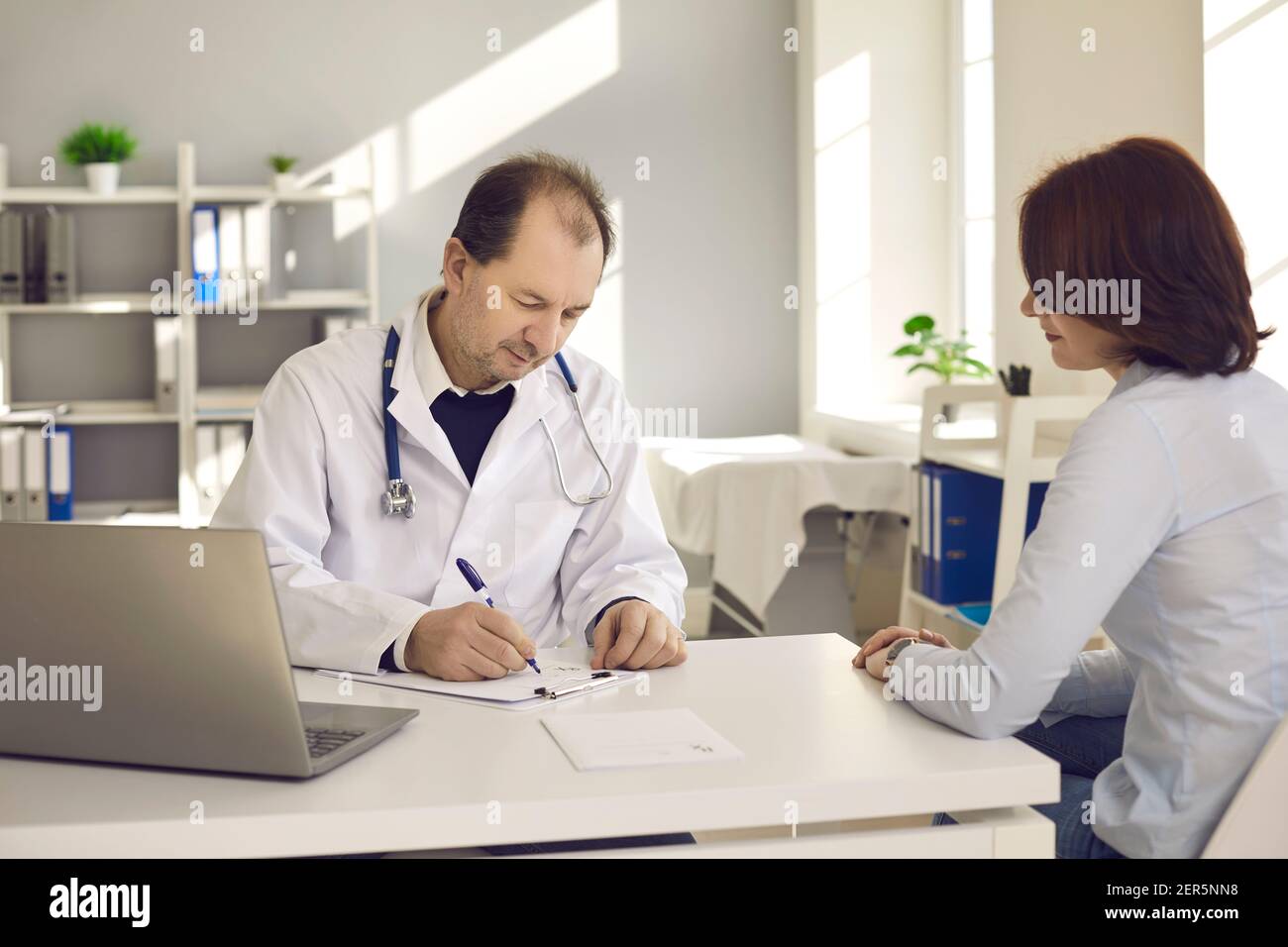 Medico di mezza età scrive una prescrizione ad una paziente femminile nel suo ufficio ospedaliero. Foto Stock