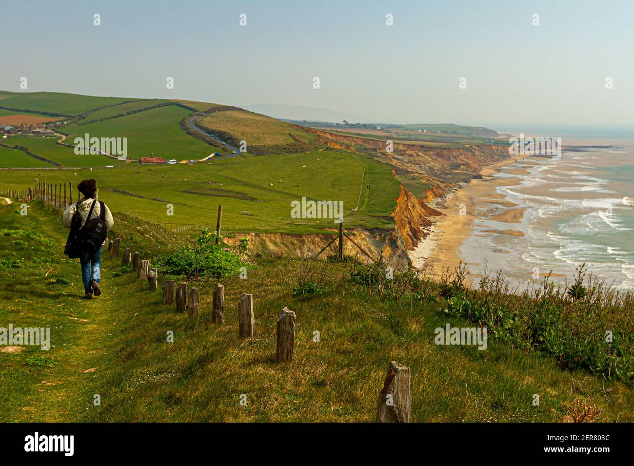 Idilliaca costa dell'Isola di Wight con scogliere, Oceano Atlantico, litorale, pascoli e fattorie a distanza e una donna escursionista con zaino a piedi da solo Foto Stock