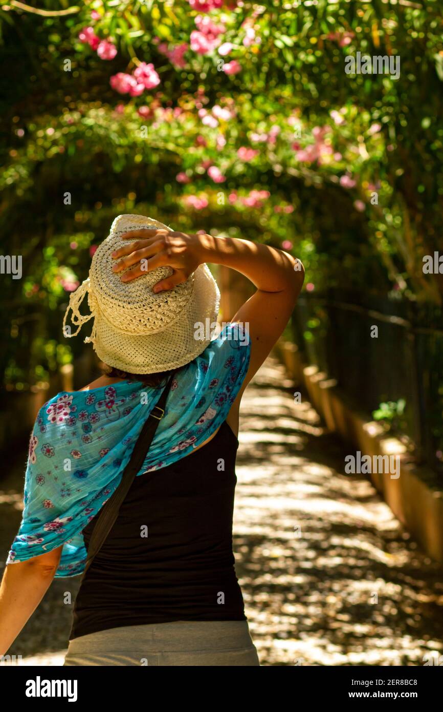 Una giovane donna caucasica che indossa un ampio cappello a cucchiaio da  sposa fatto di straffia sciolta di maglia sta camminando da solo in a.  sentiero giardino all'interno del palazzo dell'Alhambra con