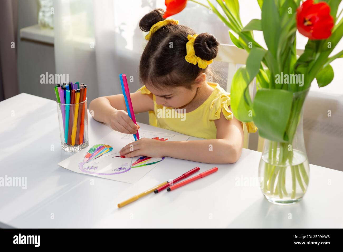 Carina bambina bruna in abito giallo, disegnando con pennarelli colorati sul tavolo bianco Foto Stock