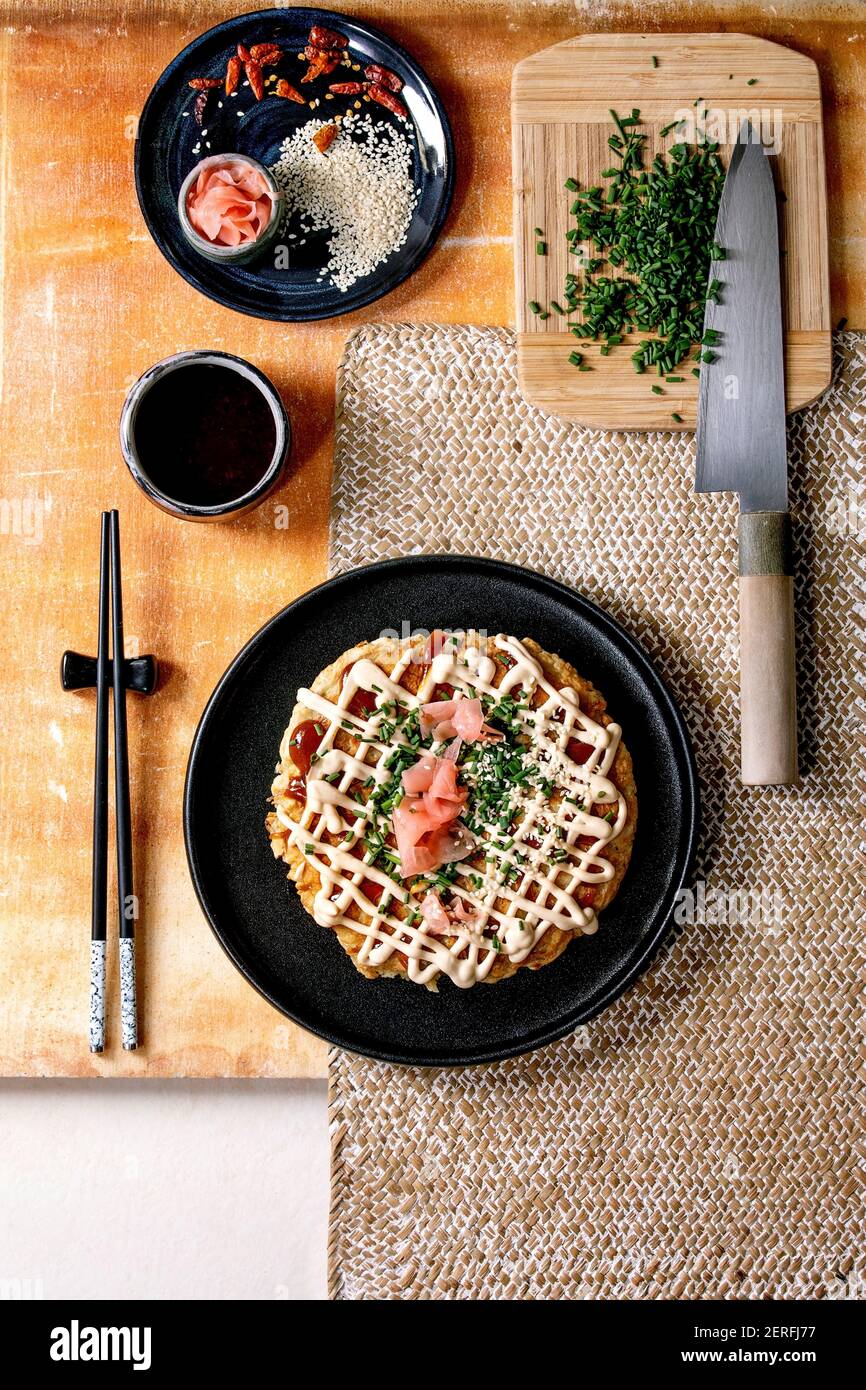 Frittella di cavolo okonomiyaki giapponese fatta in casa decorata con cipollina, zenzero sottaceto, salsa mayo su piastra di ceramica nera con chopsticks A. Foto Stock