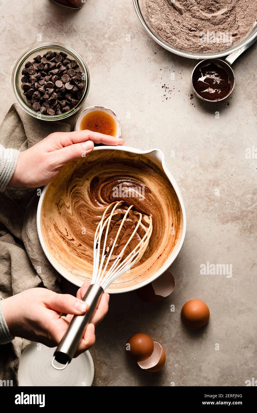 Mani che mescolano una ciotola di pastella di brownie vicino a ciotole di farina e scaglie di cioccolato. Foto Stock