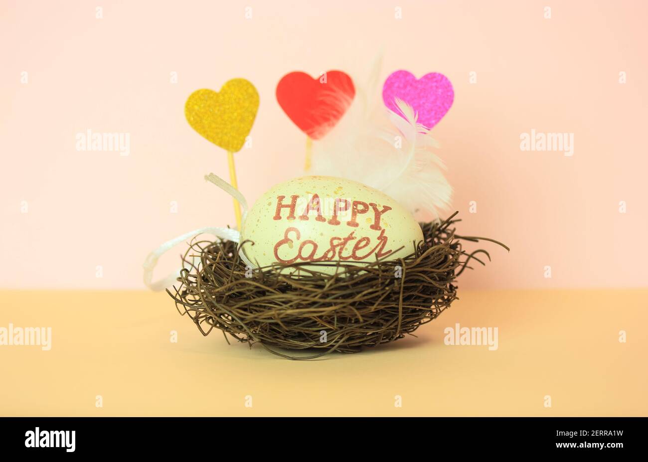 Uovo di Pasqua con Pasqua felice inscritto in un nido, cuori decorativi multicolore e una piuma bianca. Sfondo color pesca chiaro e caldo. Foto Stock