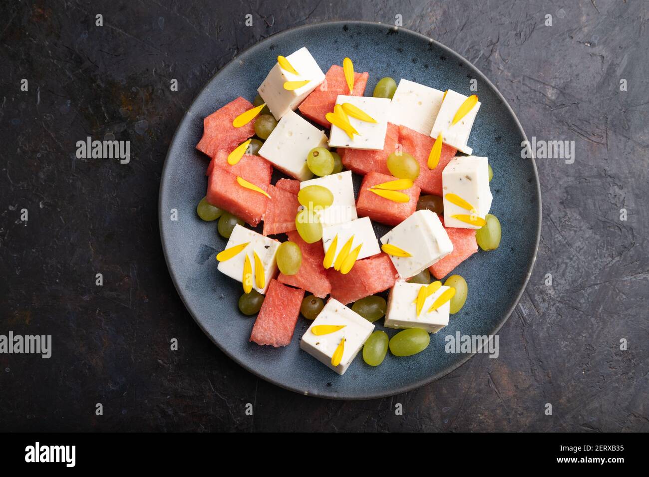 Insalata vegetariana con cocomero, formaggio feta e uva su piatto di ceramica blu su fondo di cemento nero. Vista dall'alto, primo piano, piatto. Foto Stock