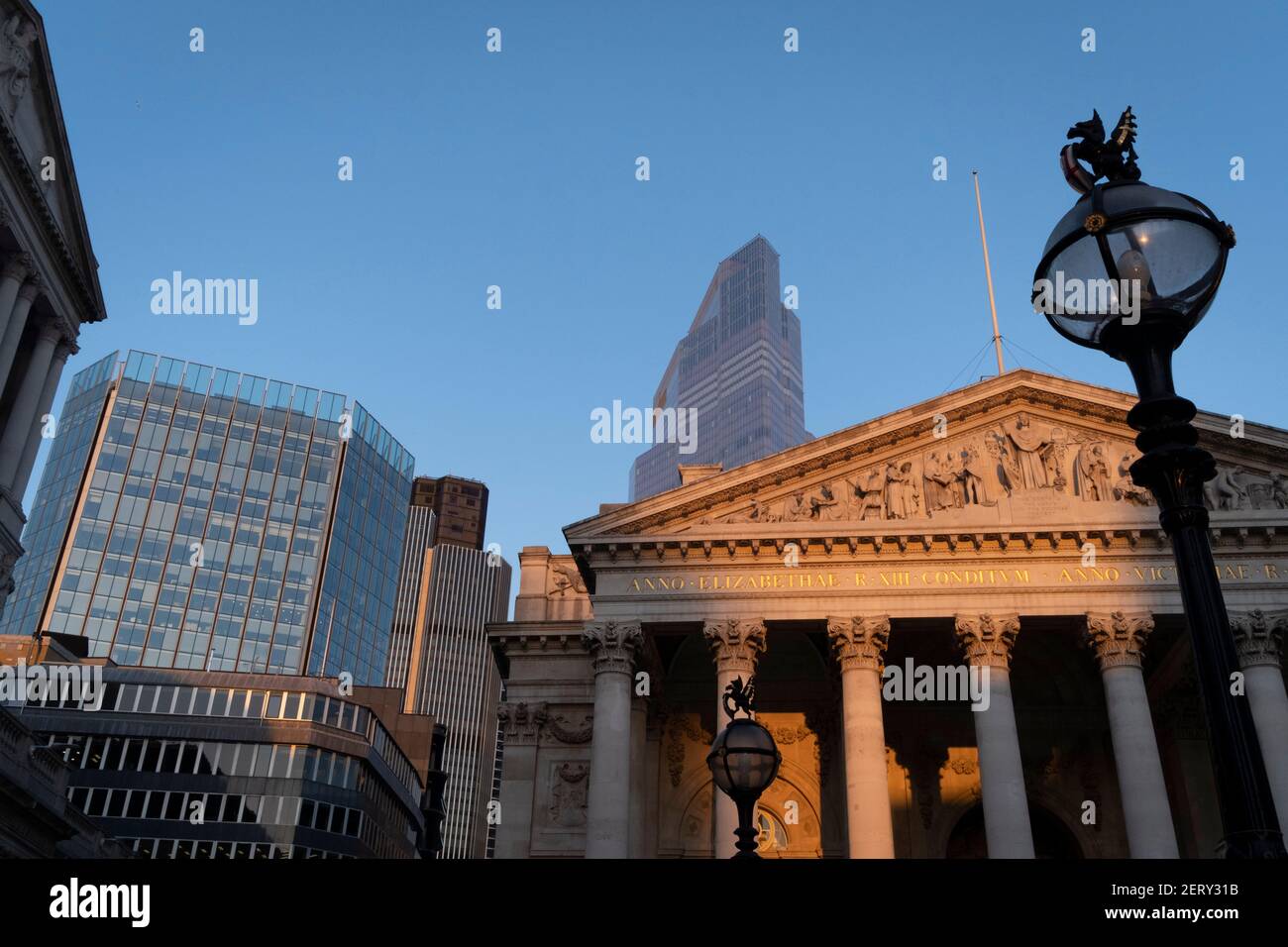 Con uffici moderni di istituzioni finanziarie dietro, una vista architettonica soleggiata dei fregi e delle iscrizioni latine sul frontone della Royal Exchange nella città di Londra, il quartiere finanziario della capitale, il 27 febbraio 2021, a Londra, Inghilterra. In cima alle colonne doriche e ioniche con le loro decorazioni in pietra, si incrociano architravi potenti, che portano il carico di belle arti e intagli che caratterizzano il disegno di Sir William Tite nel 1842-1844 e aperto nel 1844 dalla regina Vittoria il cui nome è scritto in latino (Victoriae R). E' il terzo edificio del genere eretto sulla sa Foto Stock