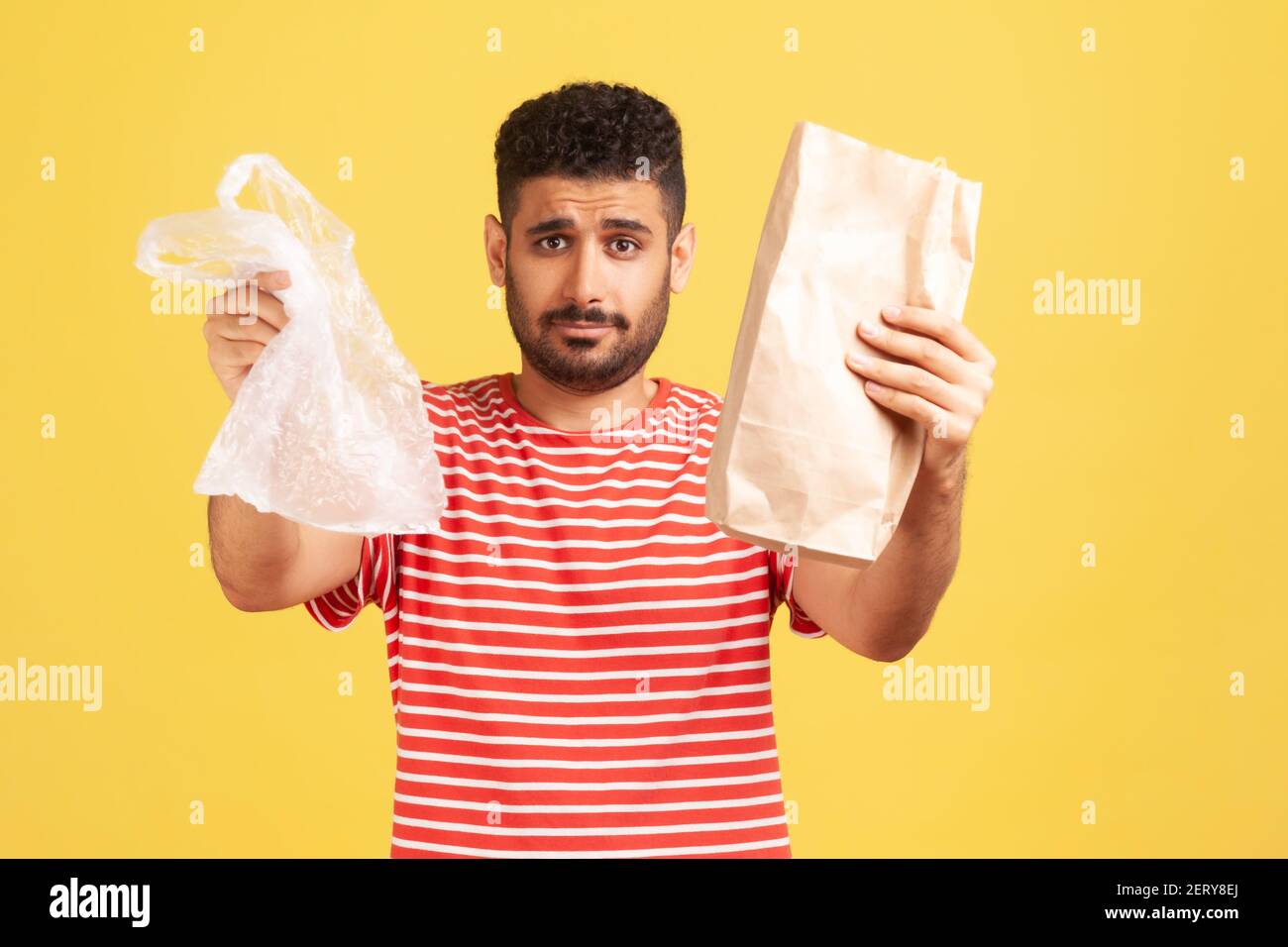 Uomo serio bearded in t-shirt a righe che tiene fuori le mani con carta e sacchetto di poly, dandogli la possibilità di fare la scelta giusta e salvare il pianeta. studi interni Foto Stock