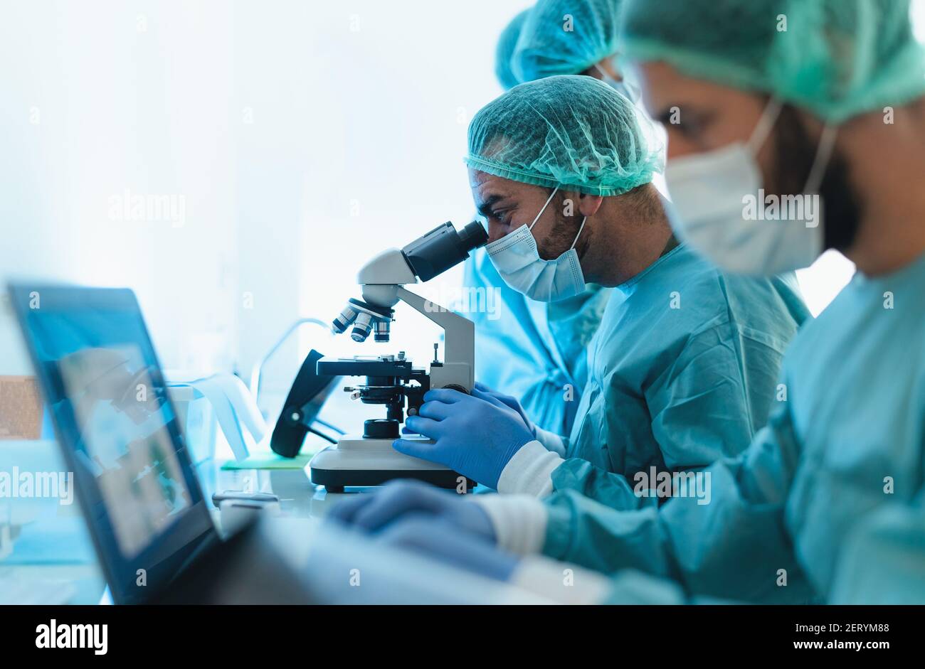 Medici che indossano dispositivi di protezione individuale che lavorano in laboratorio utilizzando il computer ed esaminare il coronavirus attraverso il microscopio Foto Stock
