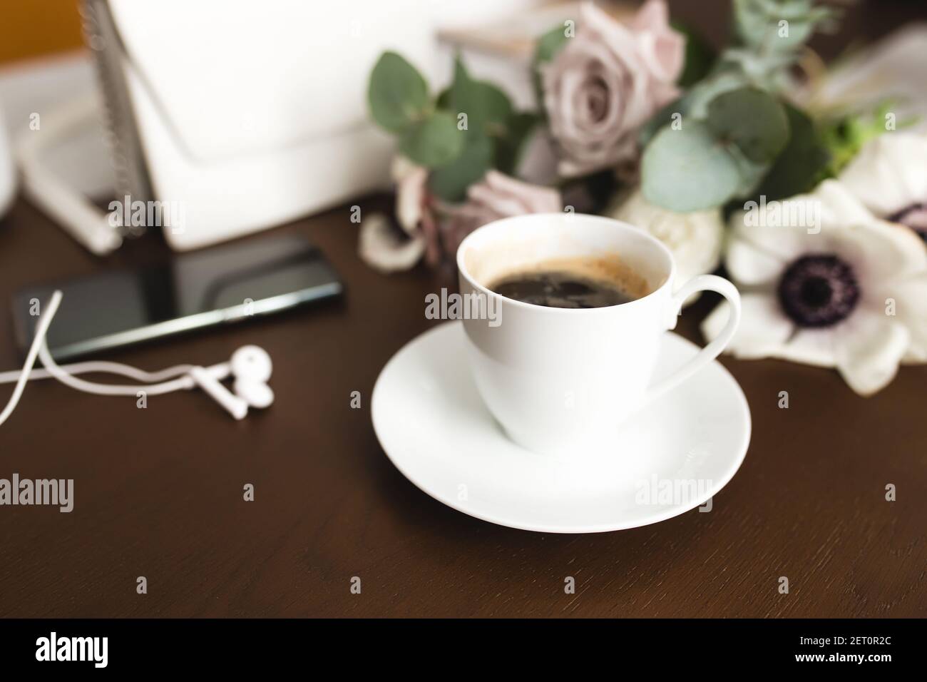 Una tazza di caffè con un bouquet di tenera anemone, rami di eucalipto e rose viola pastello, una borsa donna, cuffie e un telefono su un legno marrone Foto Stock