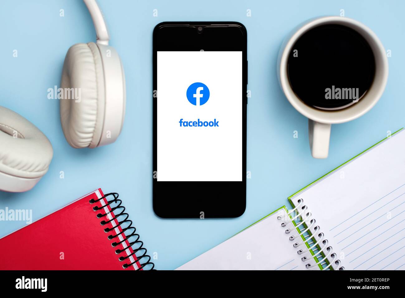 Logo Facebook sullo schermo bianco dello smartphone con notebook, cuffie e. tazza di caffè su sfondo blu Foto Stock