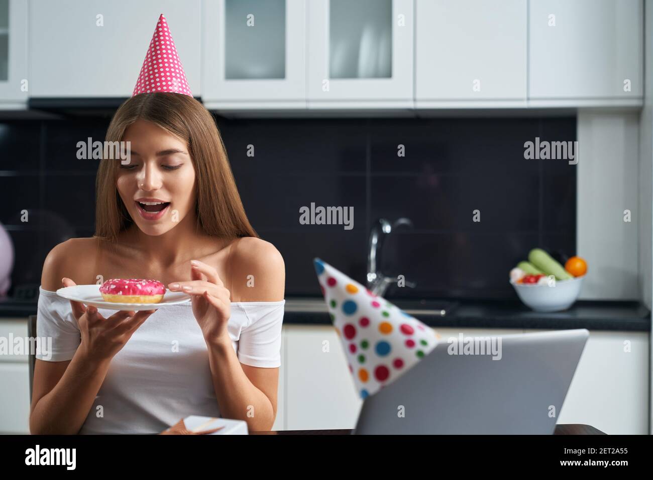 Donna allegra in cappello da festa seduto al tavolo con un portatile aperto e un piatto di tenuta con colorata e gustosa ciambella. Festa di compleanno a casa durante isola Foto Stock