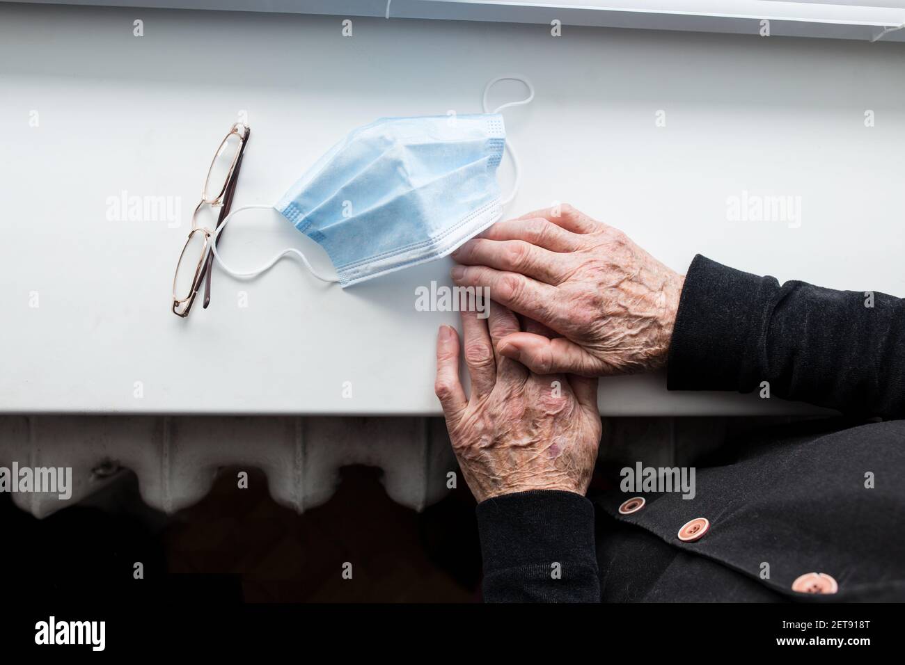 Mani di vecchio uomo con maschera medica e occhiali, accanto alla finestra. Foto Stock